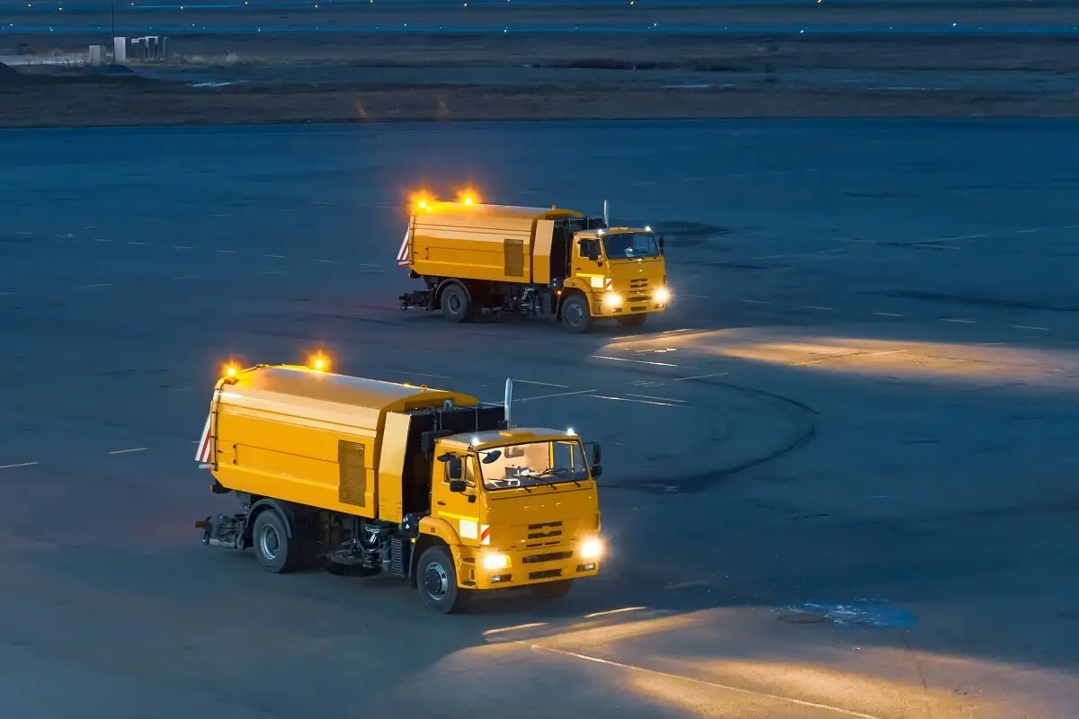 Two yellow airport runway sweepers operating at night with headlights and warning lights on.