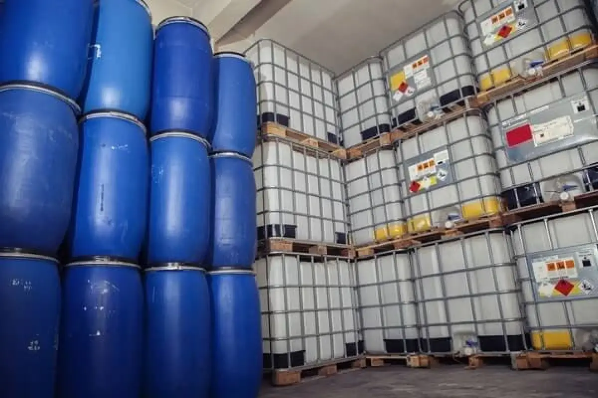 Stacks of blue industrial drums and white IBC totes stored on pallets inside a warehouse facility.