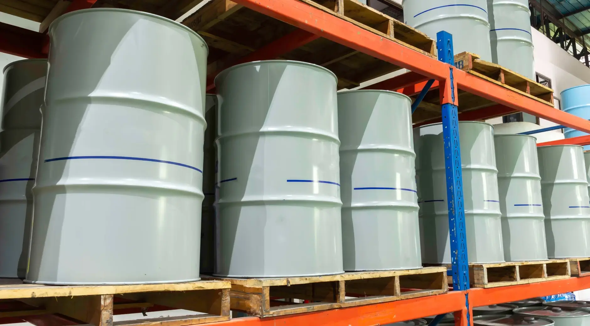 Large gray industrial storage drums stacked on wooden pallets inside a warehouse with metal shelving.